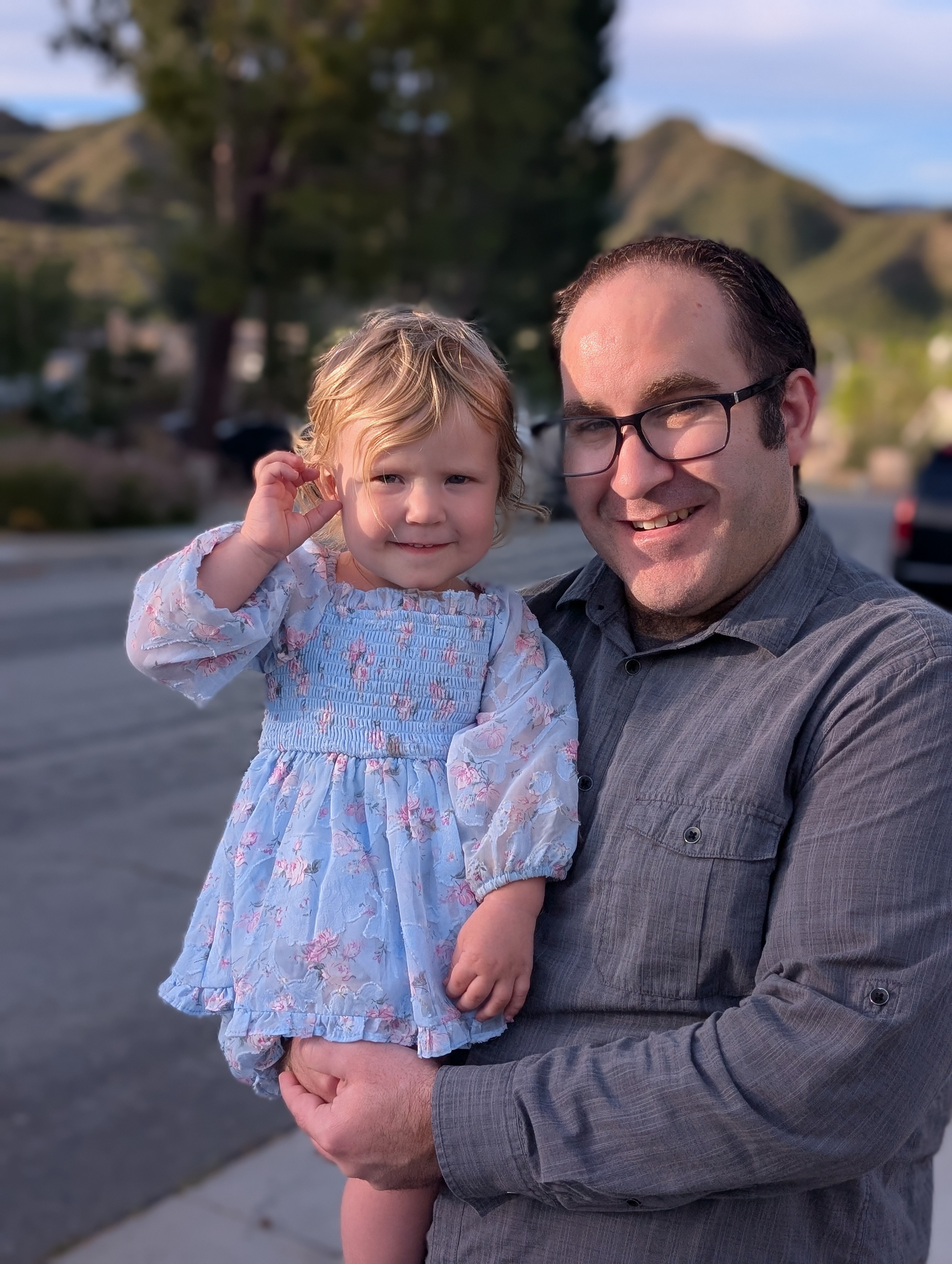 Brett Haddoak smiling and holding his young daughter, in a floral blue dress, on a neighborhood street with hills behind them.