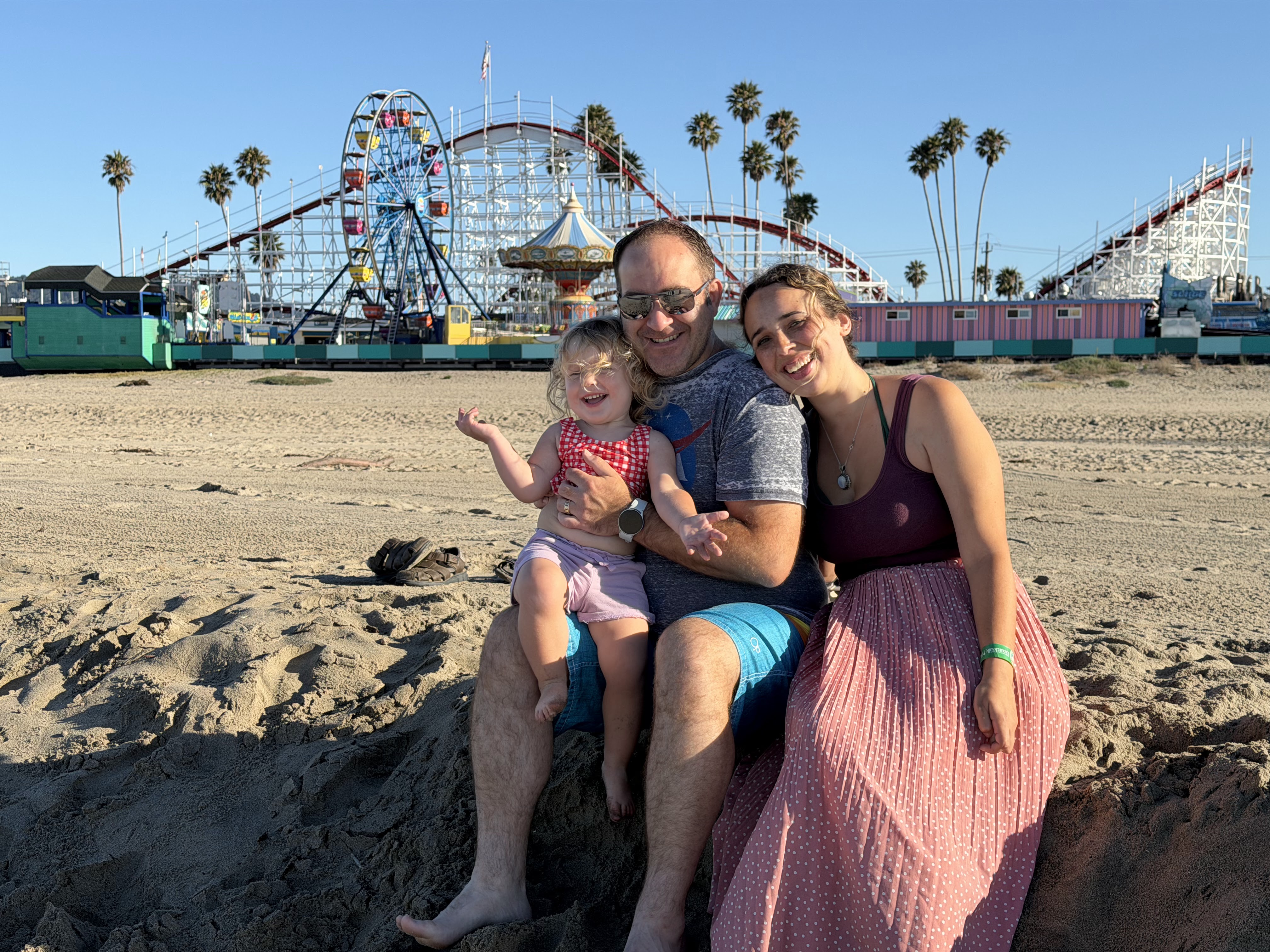 Brett Haddoak sitting on the sand with his daughter on his lap and his wife Rachelle beside him, in front of a seaside amusement park with a Ferris wheel and wooden roller coaster.