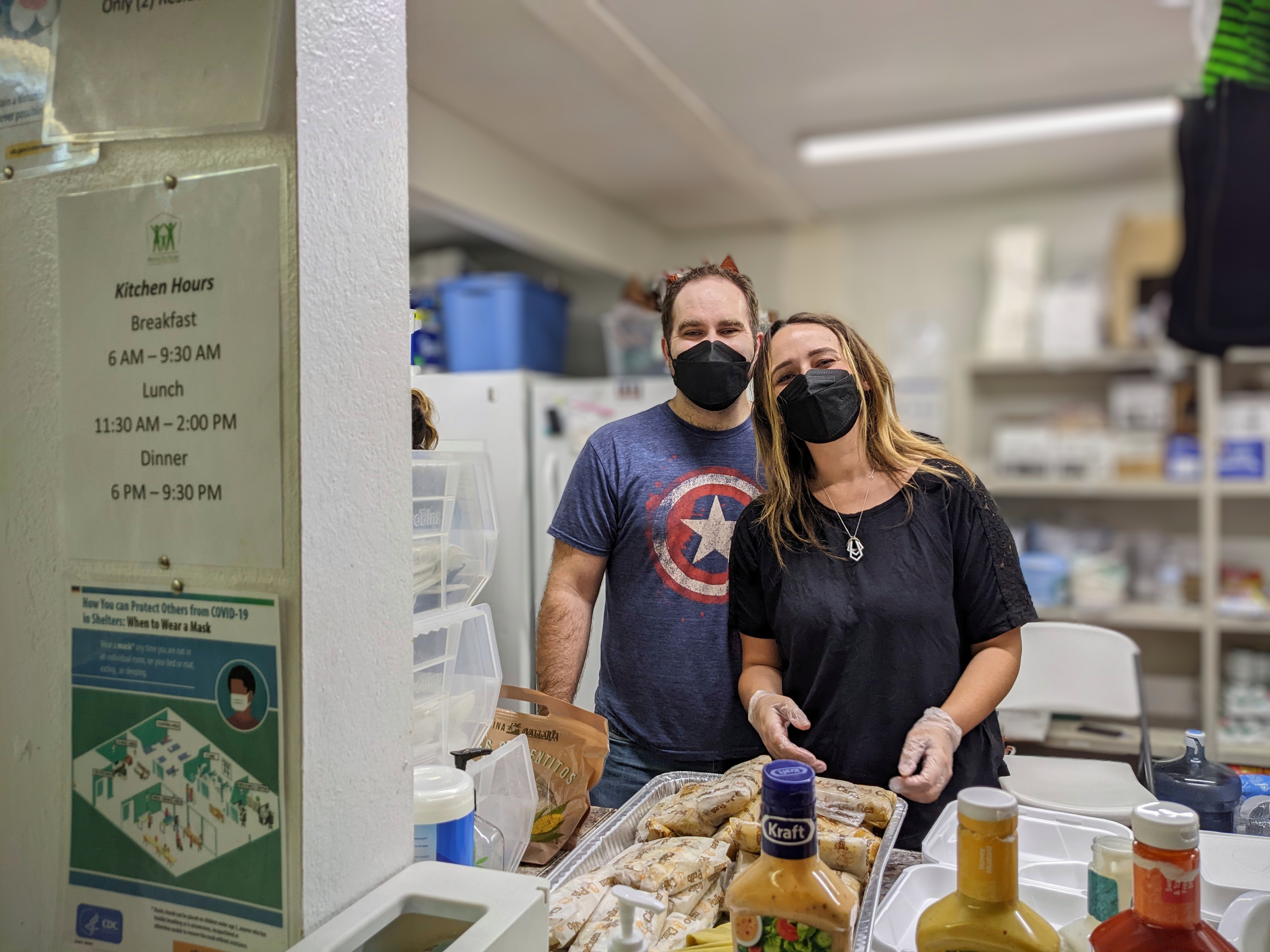Brett Haddoak and his wife Rachelle, wearing face masks and gloves, preparing a tray of food in the Bridge to Home kitchen beside a posted sign of kitchen hours.