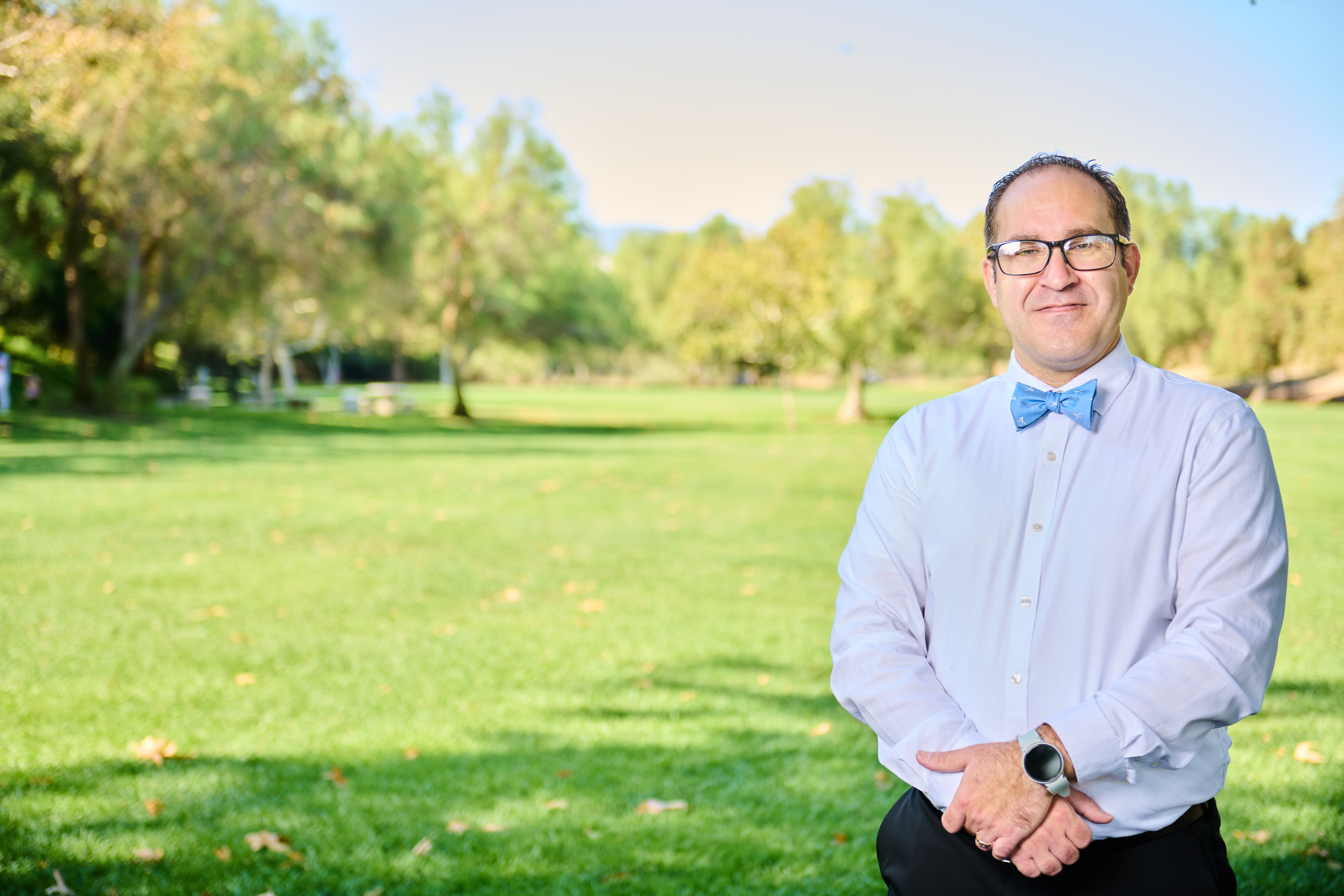 Brett Haddoak in a light blue dress shirt and blue bow tie, standing to the right of frame in a sunlit park with a broad green lawn and trees behind him.