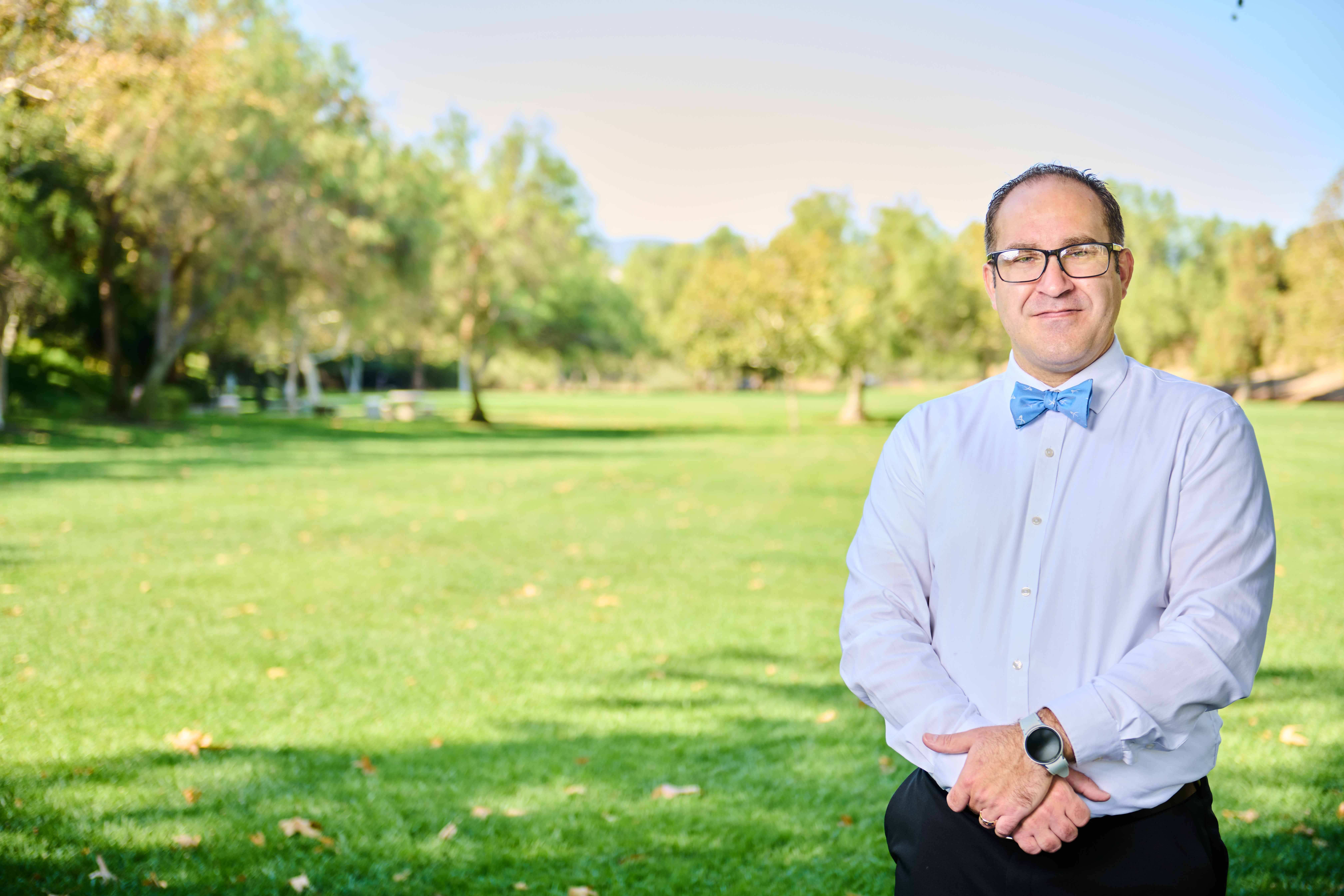Brett Haddoak in a light blue dress shirt and blue bow tie, standing to the right of frame in a sunlit park, gaze directed slightly off-camera.