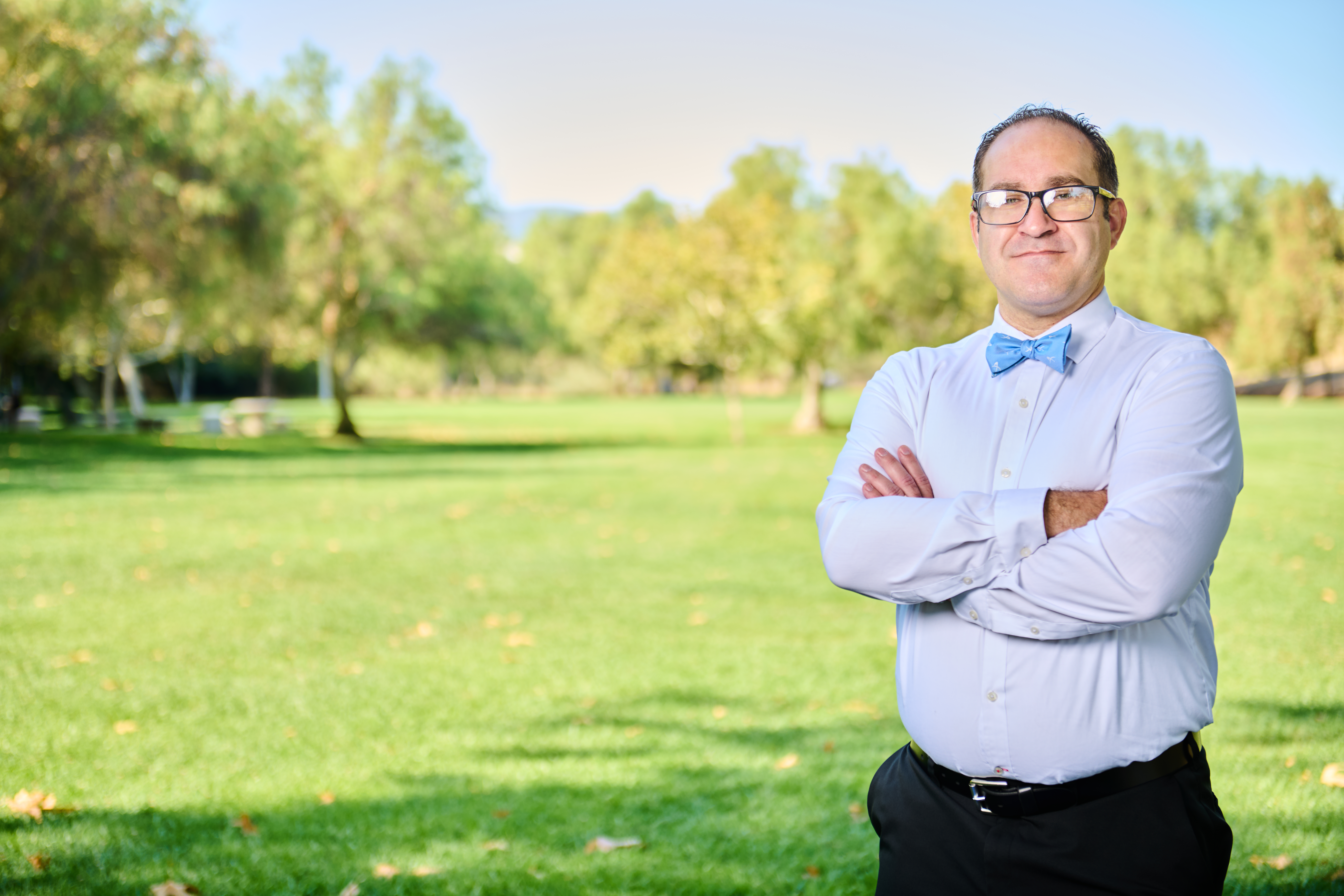 Brett Haddoak in a light blue dress shirt and blue bow tie, arms crossed, standing to the right of frame in a park with a wide green lawn.