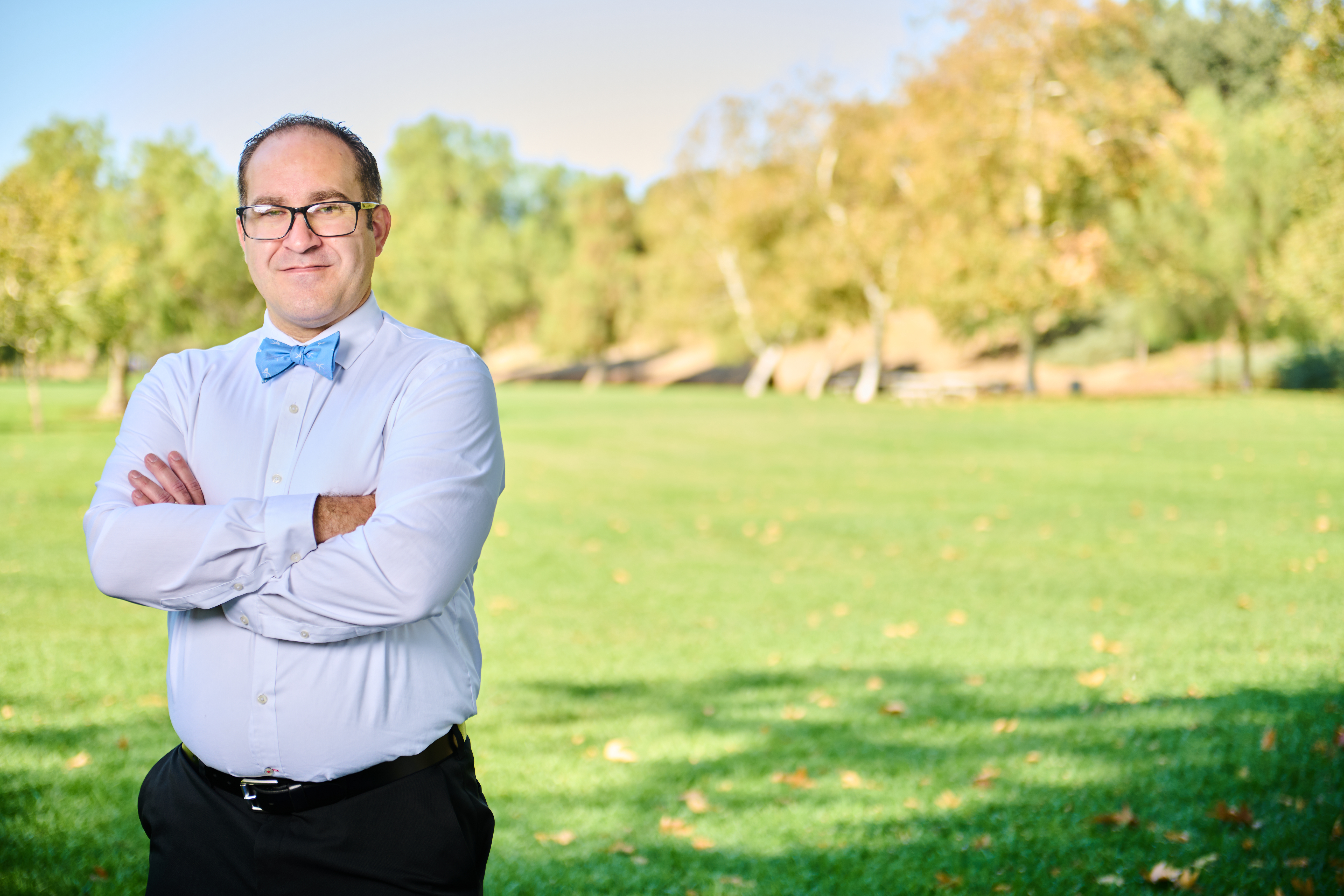 Brett Haddoak in a light blue dress shirt and blue bow tie, arms crossed, standing to the left of frame in warm late-afternoon park light.